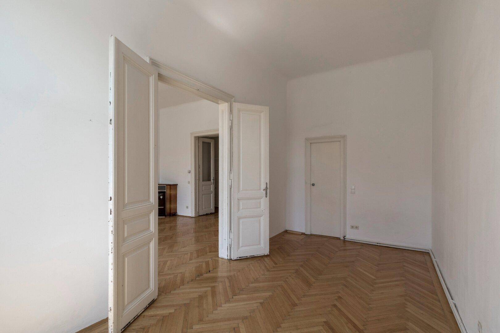 Geräumiges Zimmer mit Fischgrätparkett und hohen Flügeltüren, die den Raum öffnen.