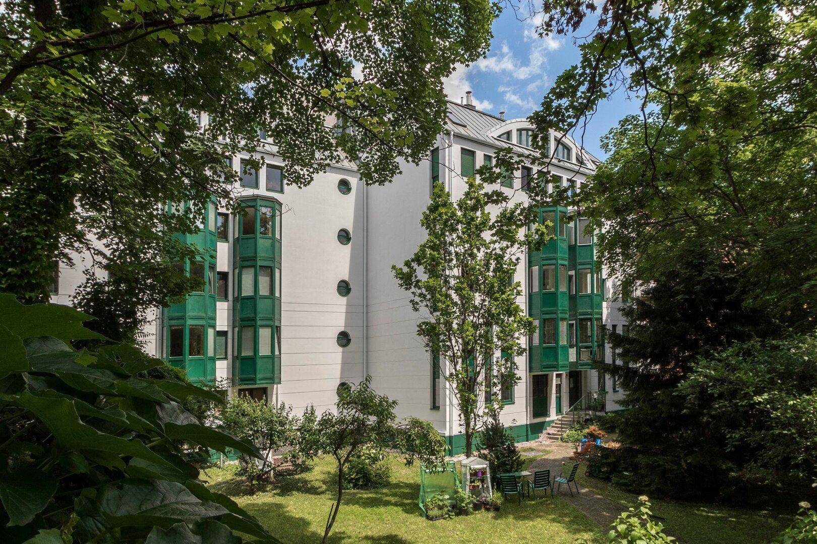 Grüne Erker und weiße Fassade eines Gebäudes, umgeben von Bäumen und einem kleinen Garten.