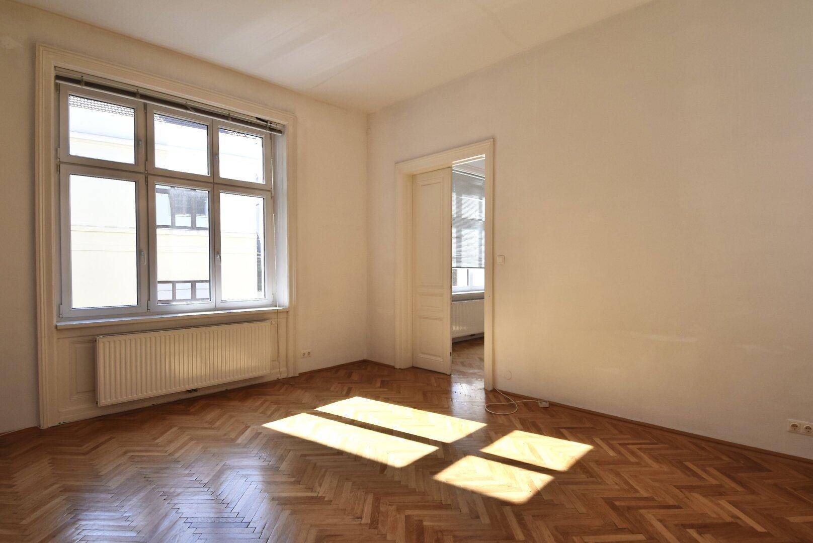 Helles Zimmer mit Fischgrätparkett und großem Fenster, das viel Sonnenlicht hereinlässt.