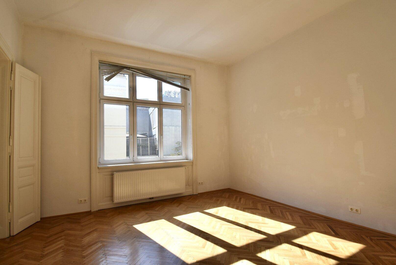 Geräumiges Zimmer mit Fischgrätparkett und viel Tageslicht durch das große Fenster.