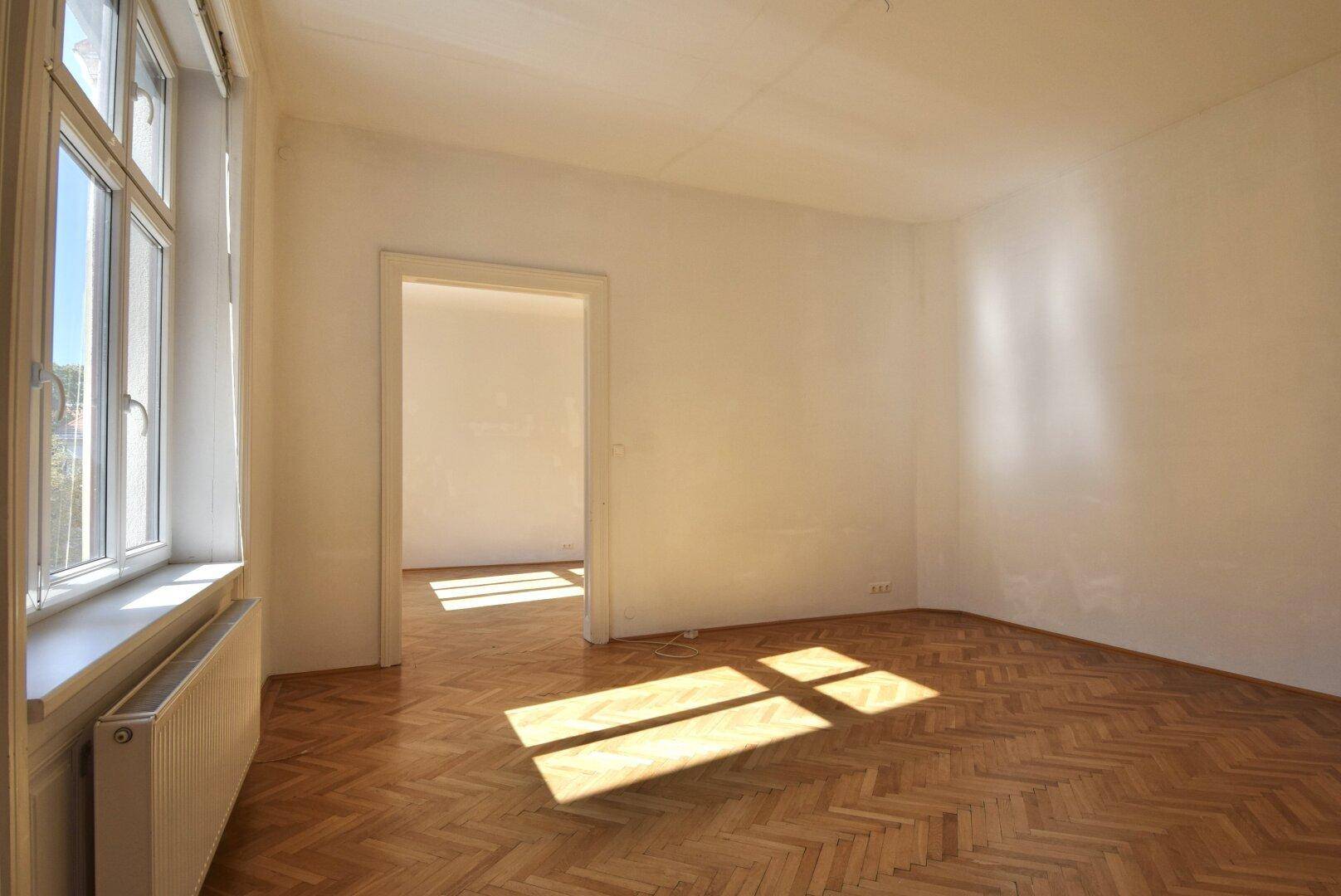 Helles Zimmer mit Fischgrätparkett und sonnigem Lichteinfall durch das Fenster.