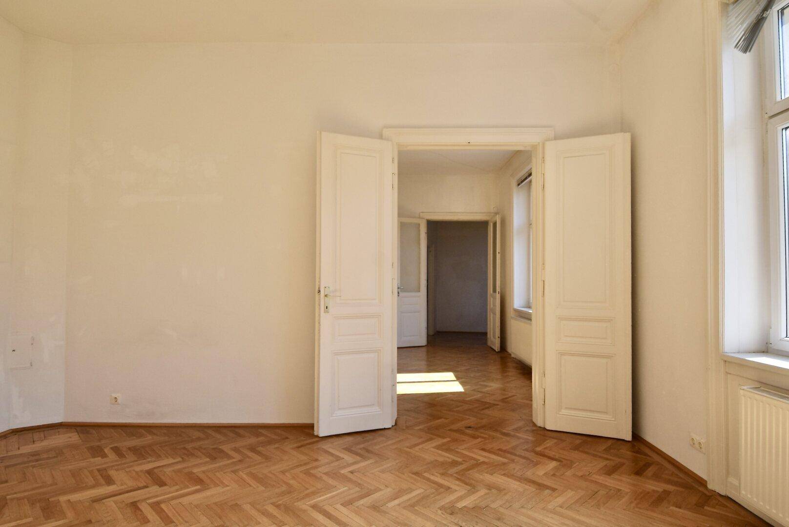 Großes Zimmer mit Fischgrätparkett und Doppeltüren, die zu weiteren Räumen führen.