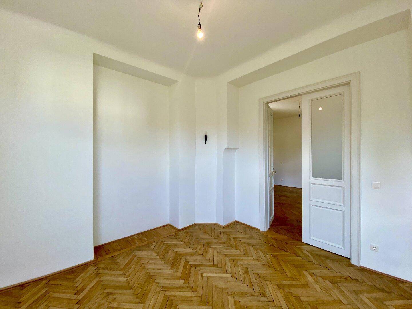 Heller Wohnraum mit Fischgrätparkett und offener Tür zu einem weiteren Zimmer.