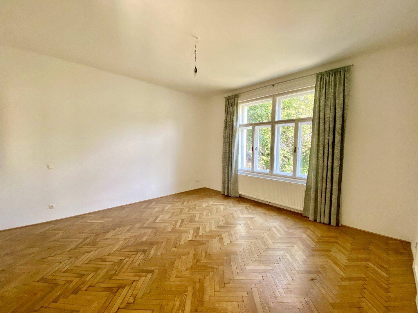 Geräumiges Wohnzimmer mit Fischgrätparkett, großen Fenstern und viel Tageslicht.