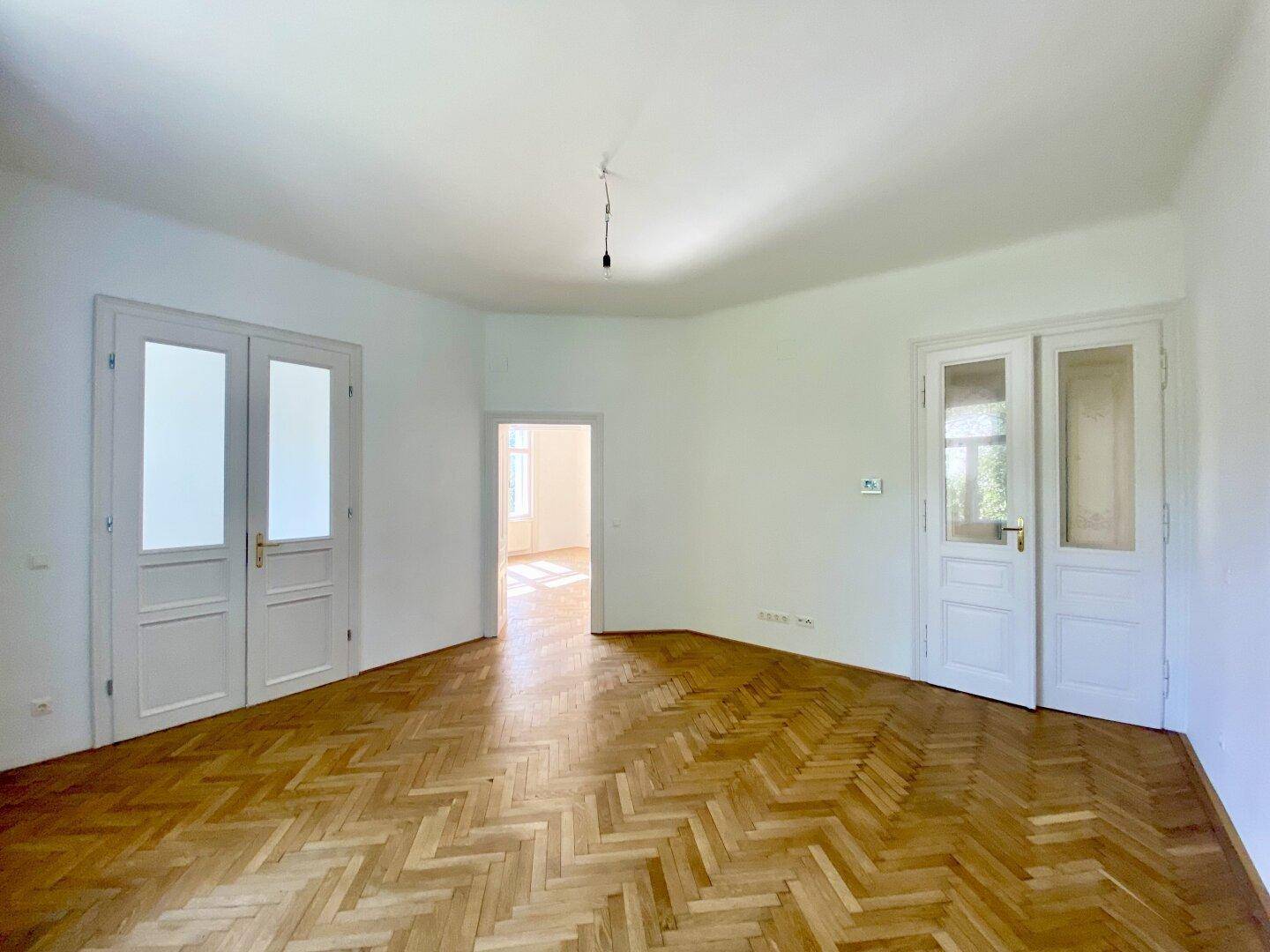 Geräumiges Wohnzimmer mit Fischgrätparkett und zwei Flügeltüren zu angrenzenden Räumen.