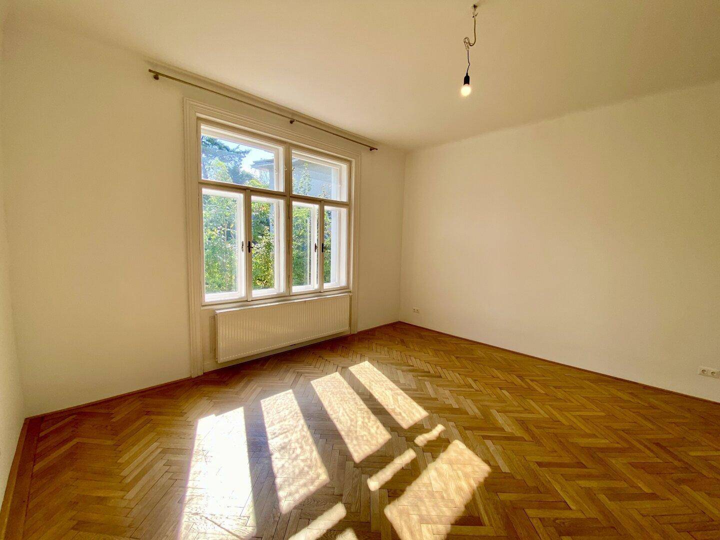 Sonniges Wohnzimmer mit Fischgrätparkett und Blick ins Grüne durch große Fenster.