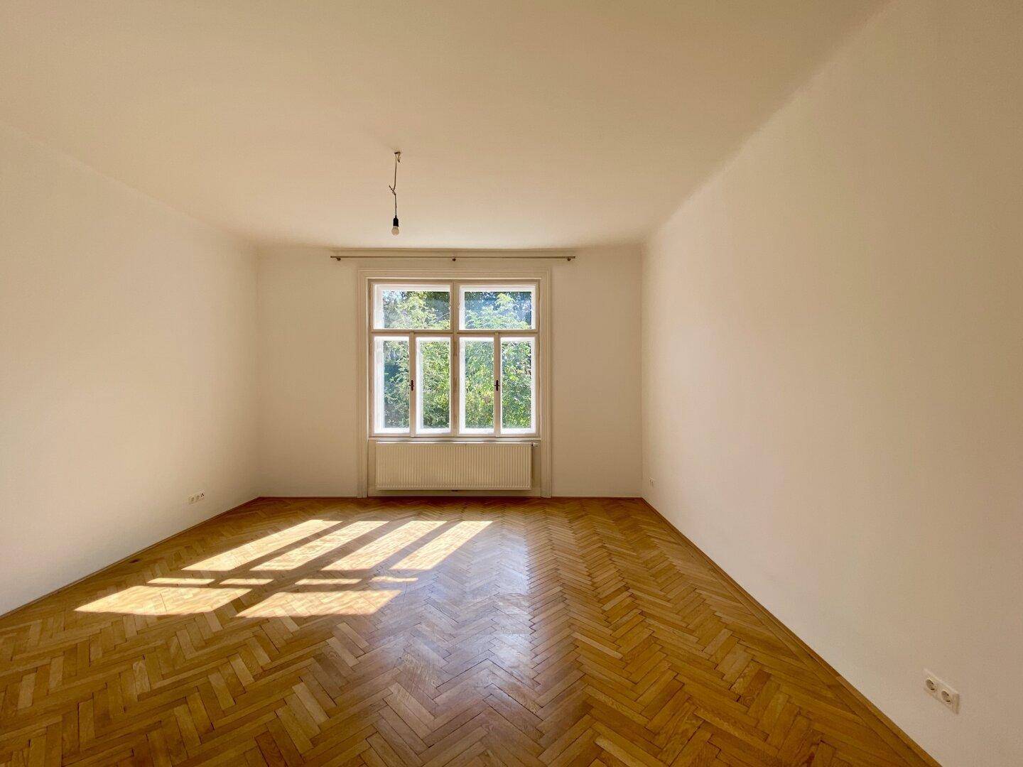 Sonniges Wohnzimmer mit Fischgrätparkett und großen Fenstern, die viel Licht hereinlassen.