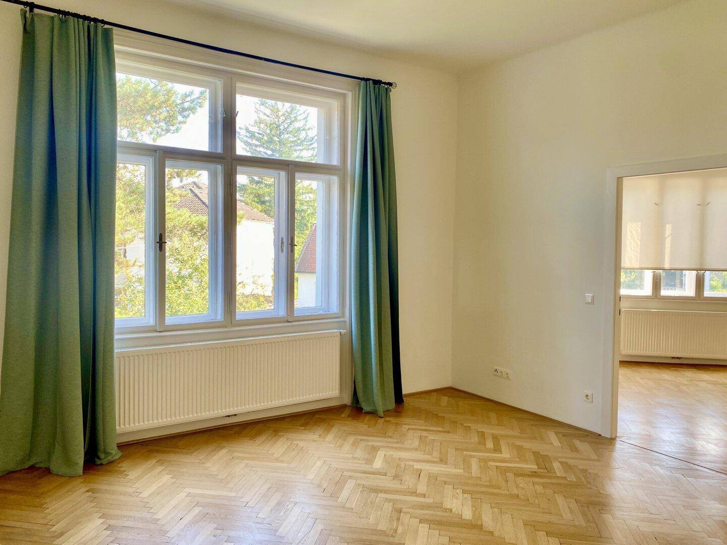 Helles Wohnzimmer mit Fischgrätparkett, großen Fenstern und grünen Vorhängen.