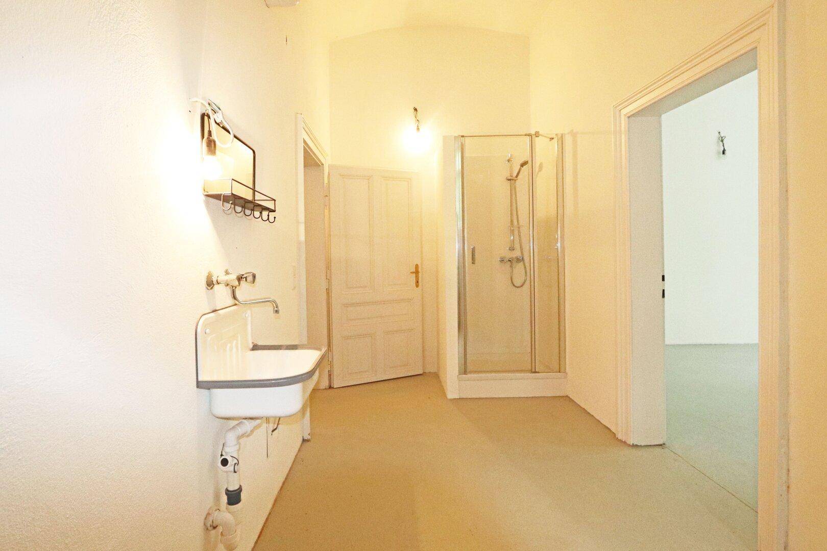 Einfaches Badezimmer mit Waschbecken, Dusche und Zugang zu weiteren Räumen.