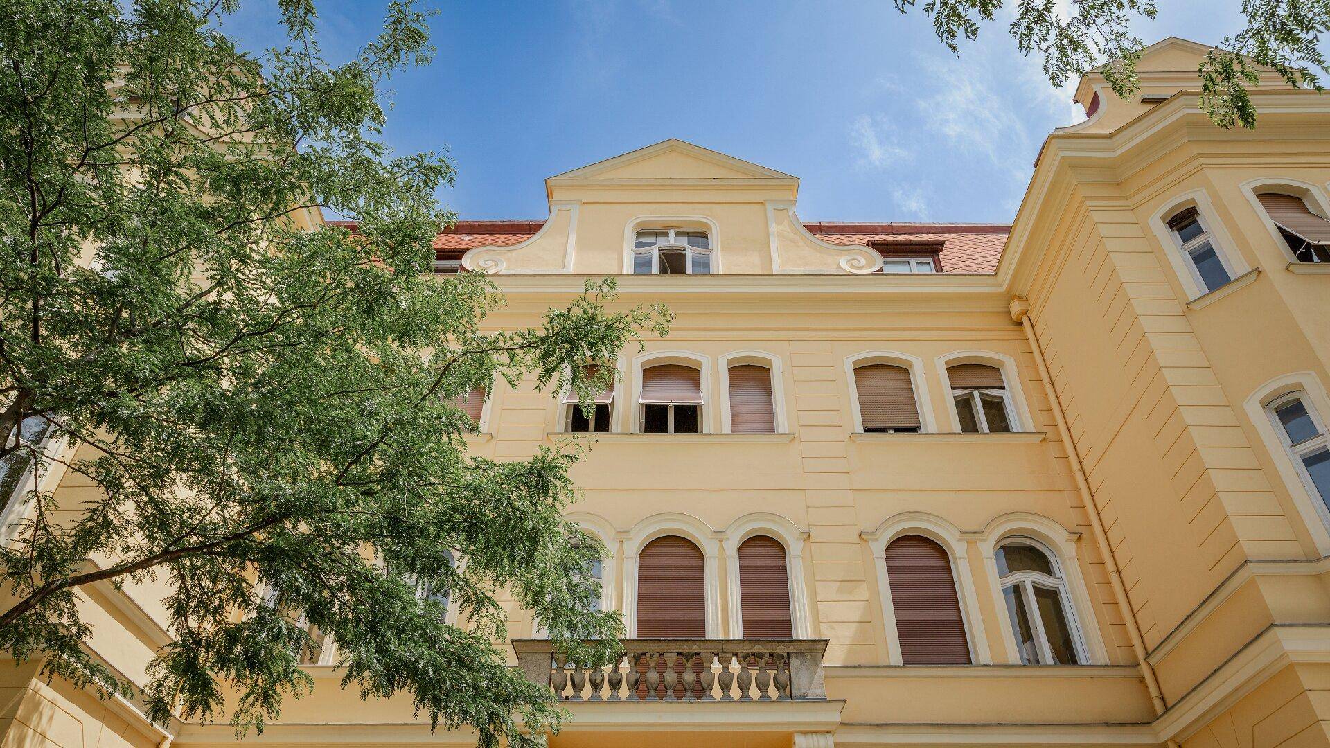 Detailansicht der gelben Fassade mit klassischen Fenstern, Rollläden und einem kleinen Balkon.