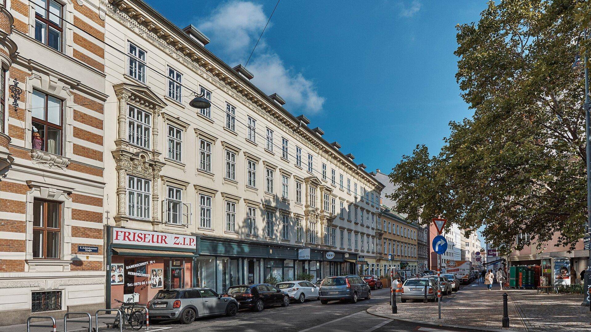 Historische Straße mit Gebäuden, Geschäften und parkenden Autos unter blauem Himmel.