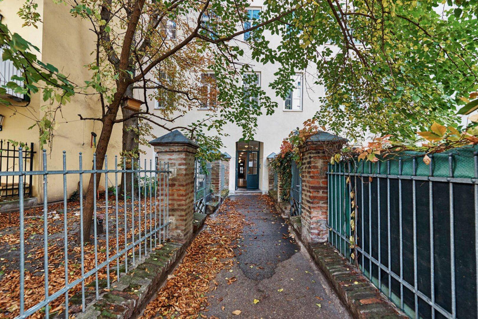 Zugang zum Gebäude durch einen begrünten Weg mit Herbstlaub und schmiedeeisernem Zaun.