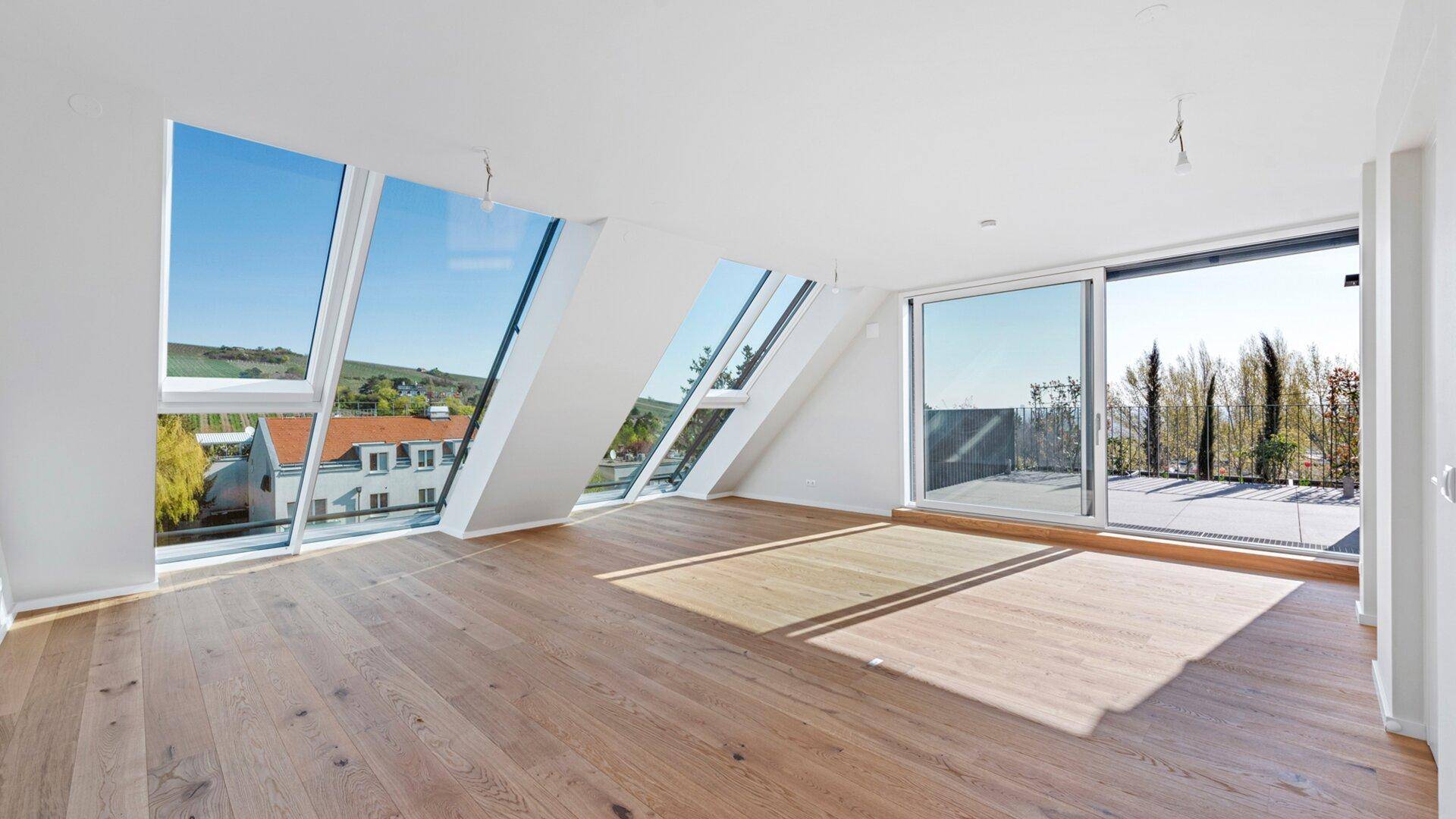 Helles Wohnzimmer mit großen Schrägfenstern, Holzboden und Zugang zu einer sonnigen Terrasse.