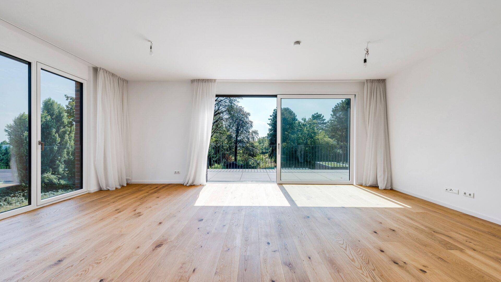 Helles Wohnzimmer mit großen Fenstern, weißen Vorhängen, Holzboden und Blick ins Grüne.