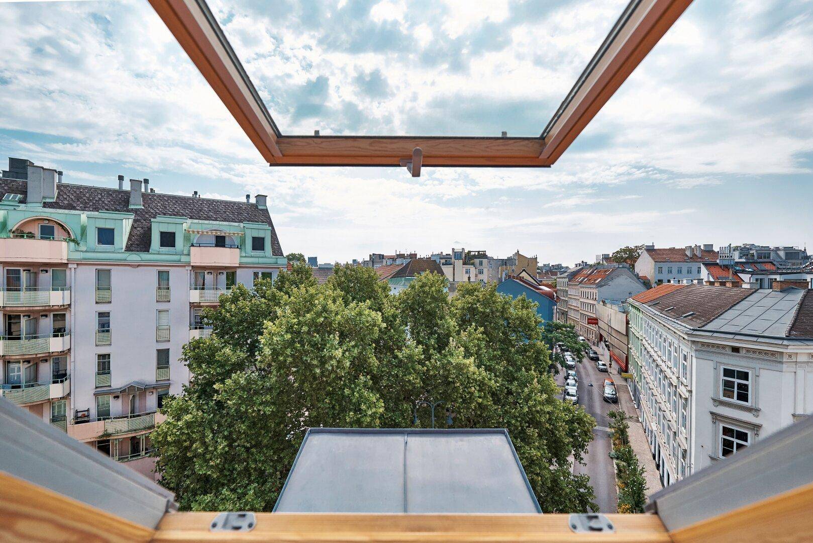 Blick aus einem Dachfenster auf die Stadtlandschaft mit Gebäuden und Bäumen unter bewölktem Himmel.