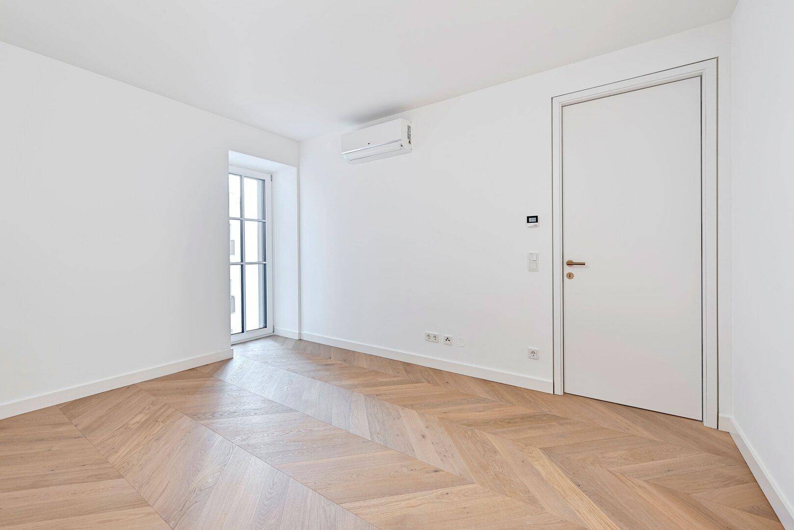 Helles Schlafzimmer mit Fischgrätparkett, einem hohen Fenster und Klimaanlage.