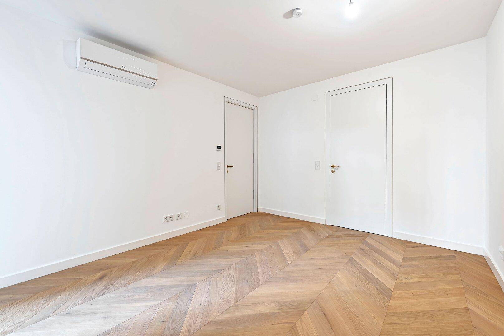 Leeres Schlafzimmer mit Fischgrätparkett, weißen Wänden und Klimaanlage.