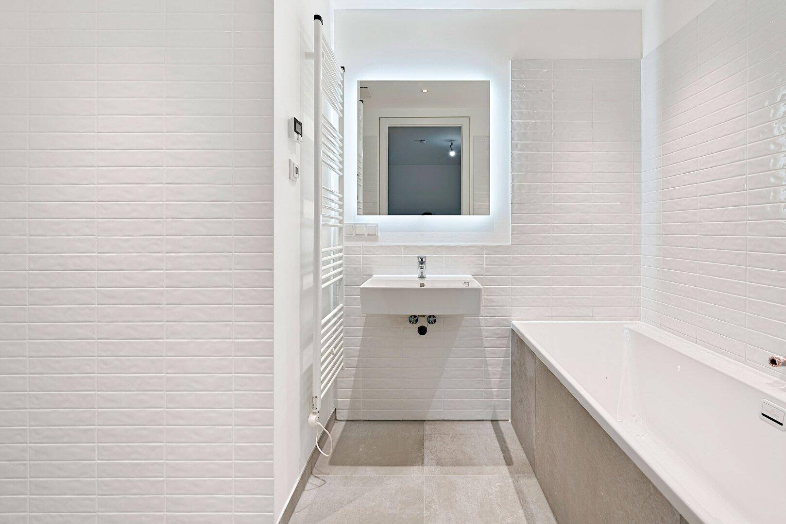 Modernes Badezimmer mit weißen Fliesen, Badewanne, Einzelwaschbecken und beheiztem Handtuchhalter.
