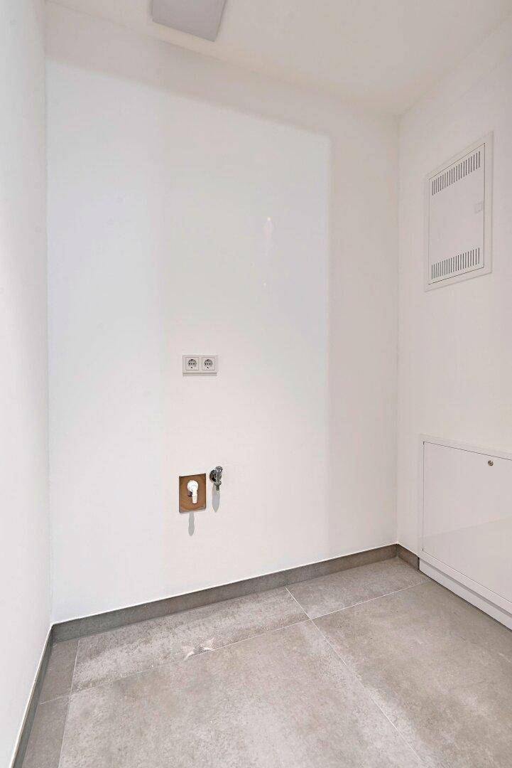 Leerer Hauswirtschaftsraum mit grauen Bodenfliesen und Anschlüssen für Waschmaschine oder Trockner.