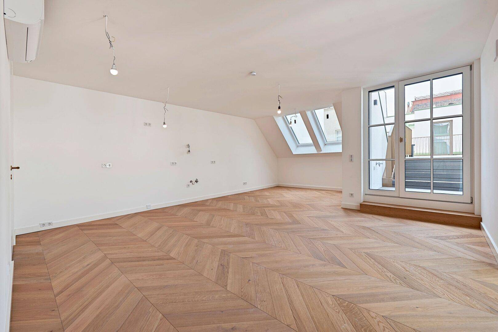 Lichtdurchflutetes Zimmer mit Fischgrätparkett, Dachfenstern und Zugang zu einem französischen Balkon.