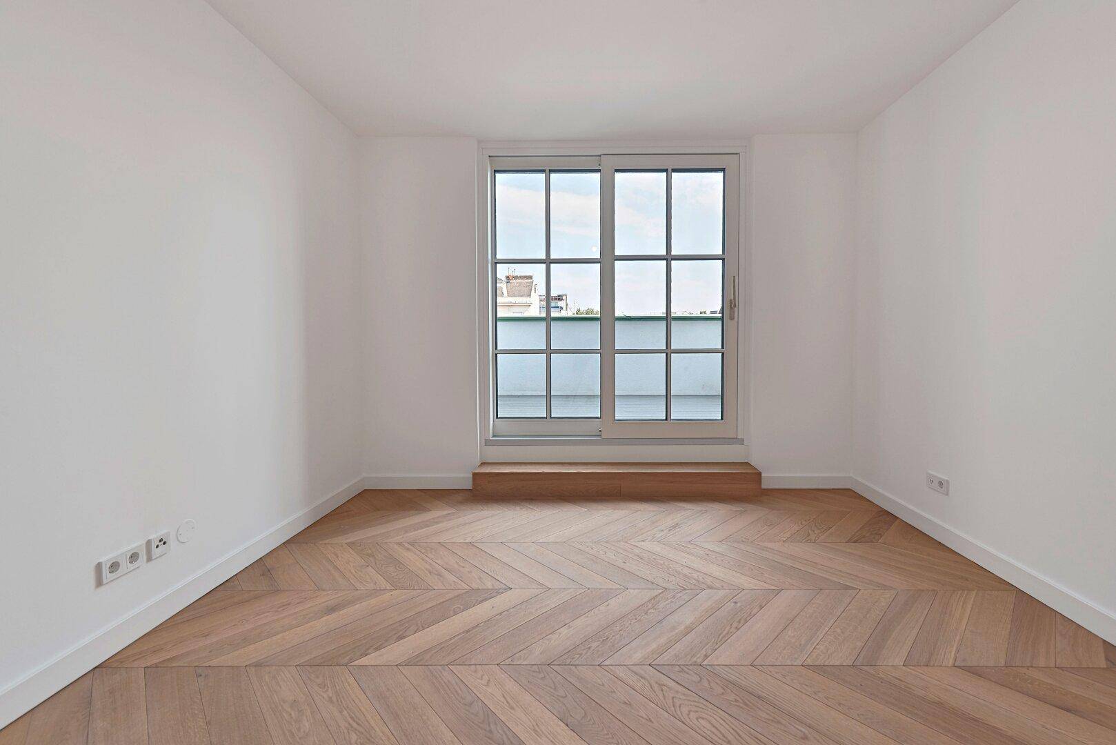 Helles, leeres Zimmer mit Fischgrätparkett und großem Fenster mit Blick nach draußen.