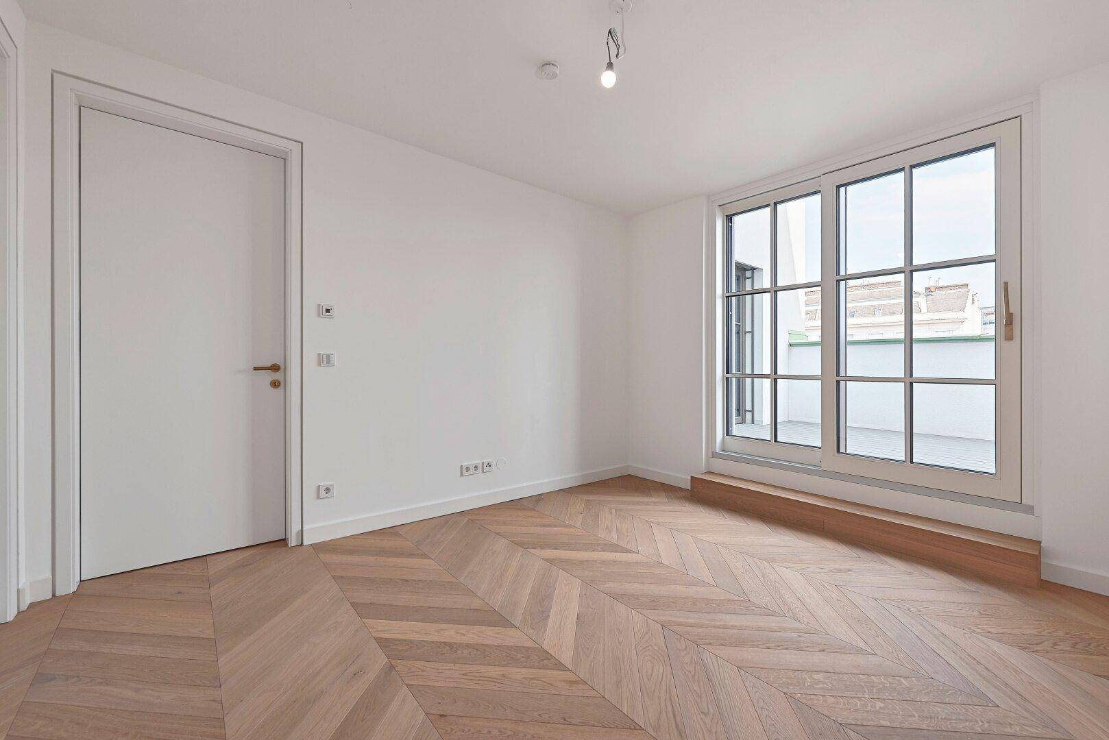 Helles Zimmer mit Fischgrätparkett, moderner Tür und Zugang zu einem französischen Balkon.