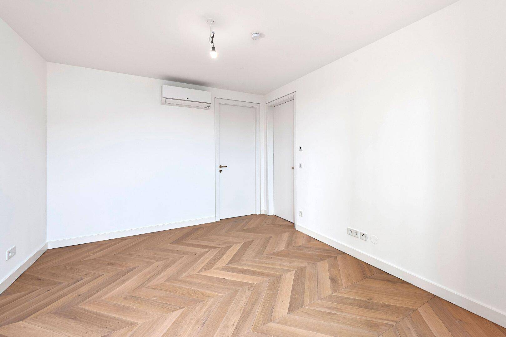 Geräumiges, helles Zimmer mit Fischgrätparkett, Klimaanlage und moderner weißer Tür.