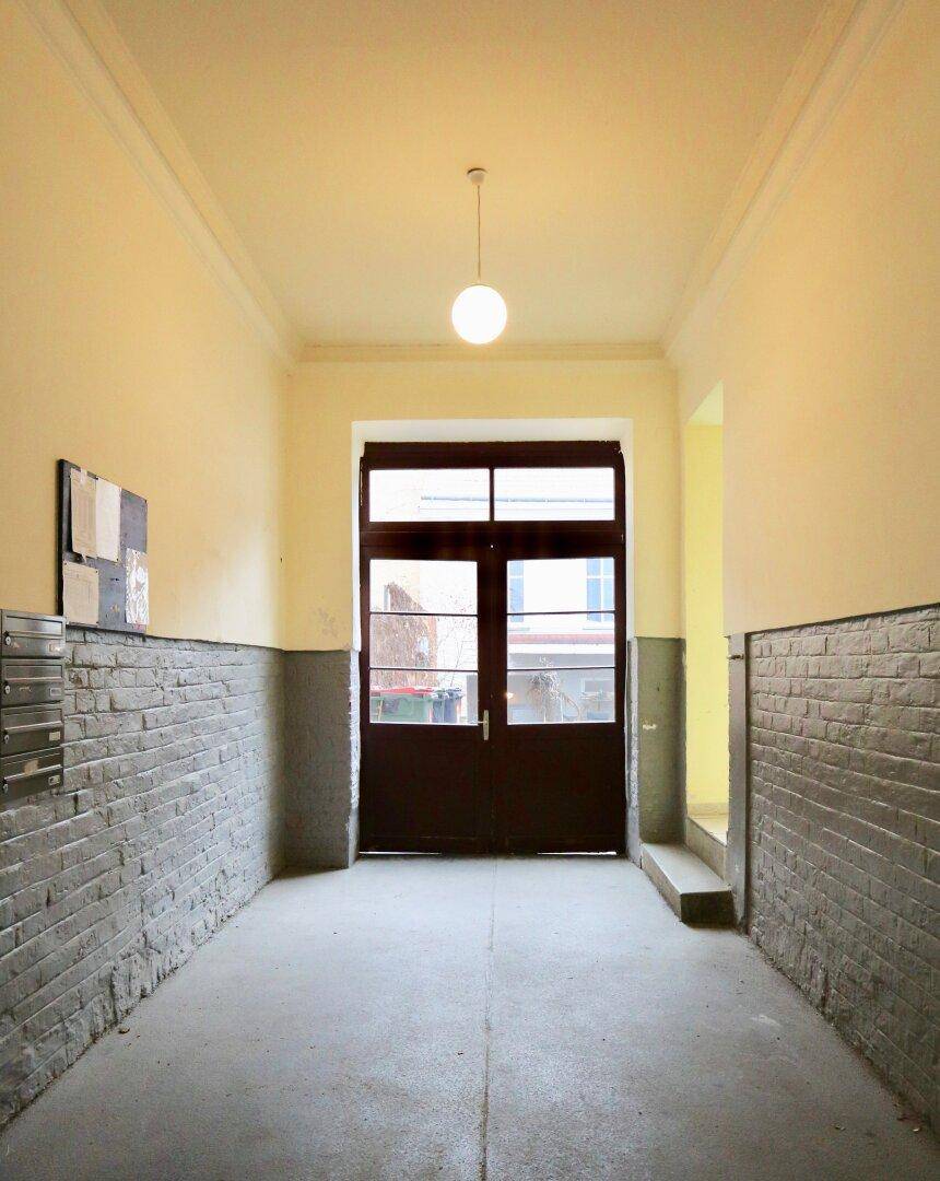 Langer Flur in einem Altbau mit gelben Wänden, Ziegelverkleidung und einer großen Eingangstür.