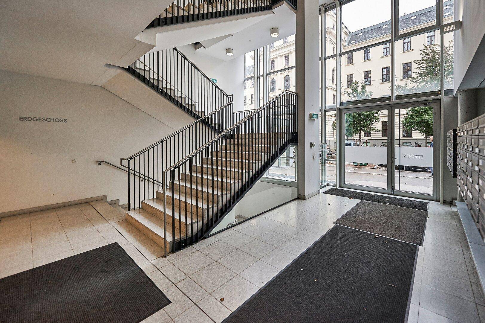 Heller Eingangsbereich mit breiter Treppe, Fliesenboden und großen Fenstern zur Straße.