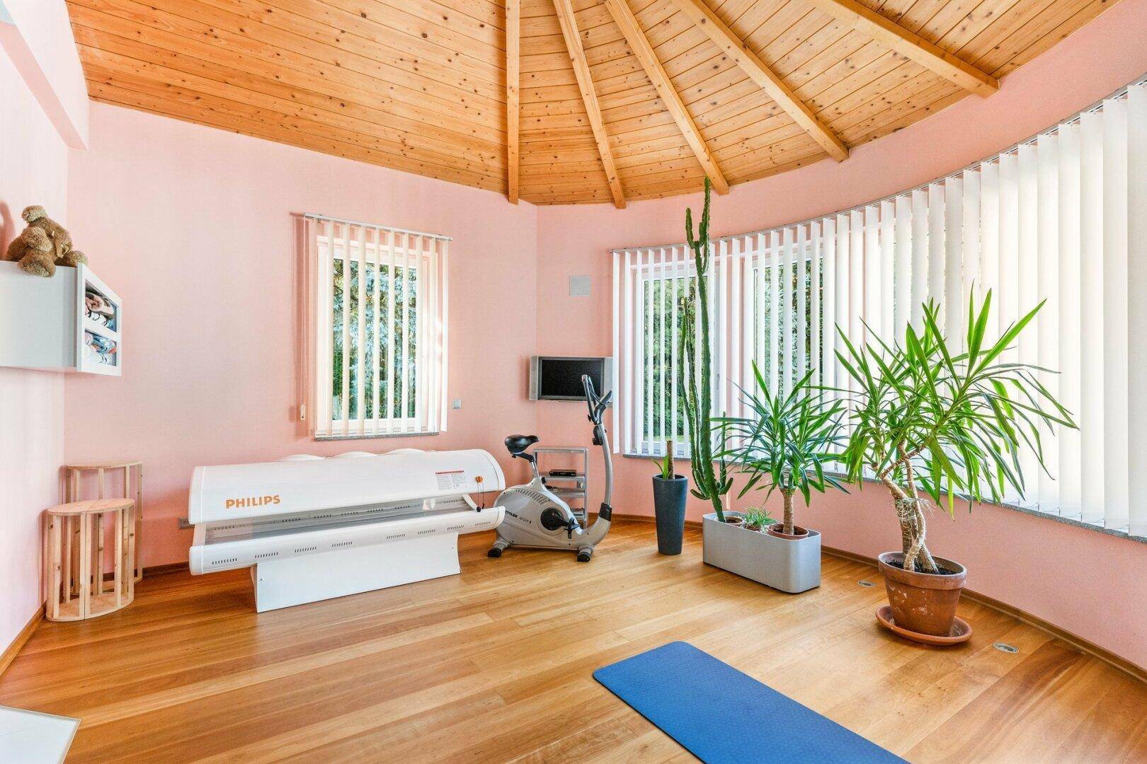 Fitnessraum mit Solarium, Trainingsgerät und Holzboden unter einer Holzdecke.