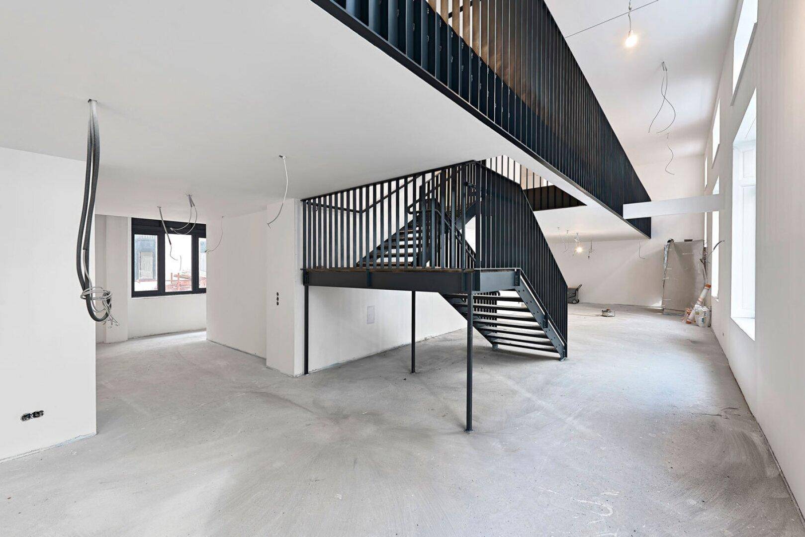 Unfertiger Büroraum mit offener Galerie, schwarzer Metalltreppe und Betonboden, bereit für den Ausbau.