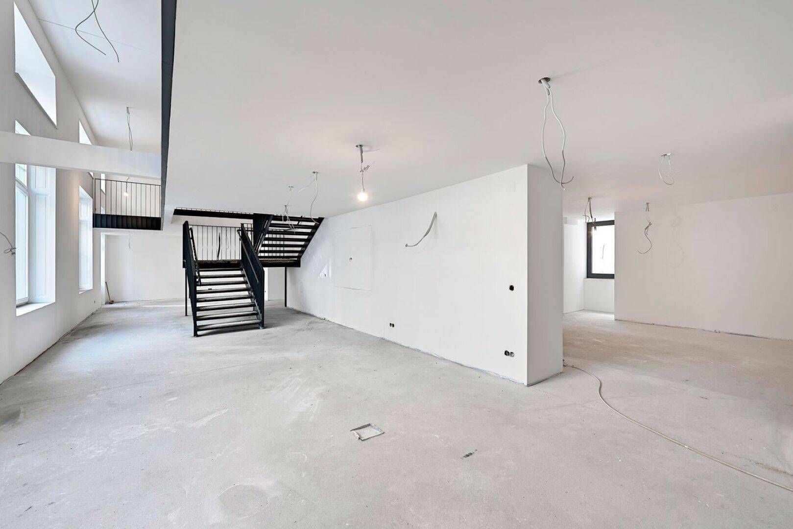 Großer, leerer Büroraum im Rohbauzustand mit Betonboden und freiliegenden Kabeln an der Decke.