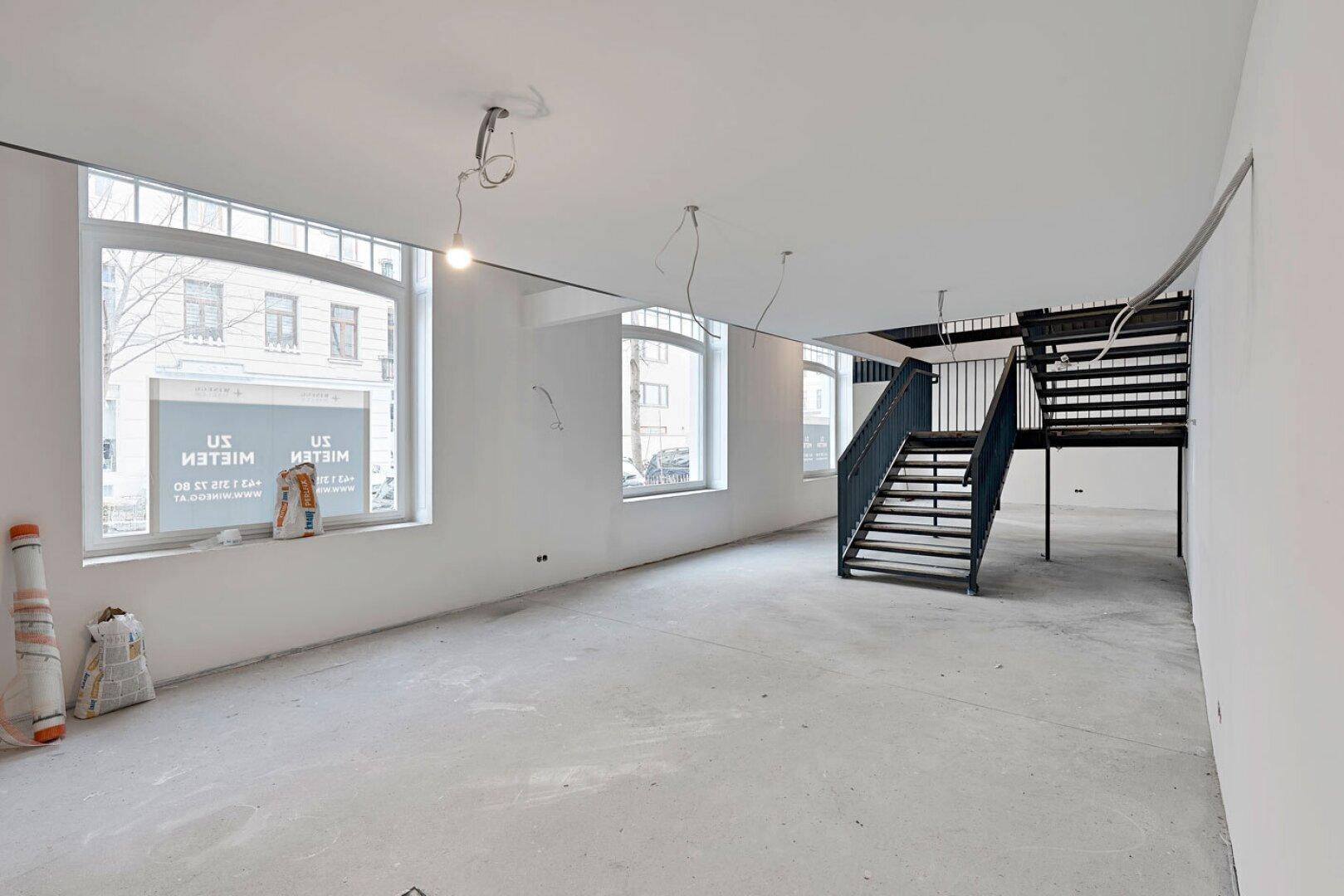 Heller Büroraum im Rohbau mit großen Fenstern zur Straße und einer schwarzen Metalltreppe.