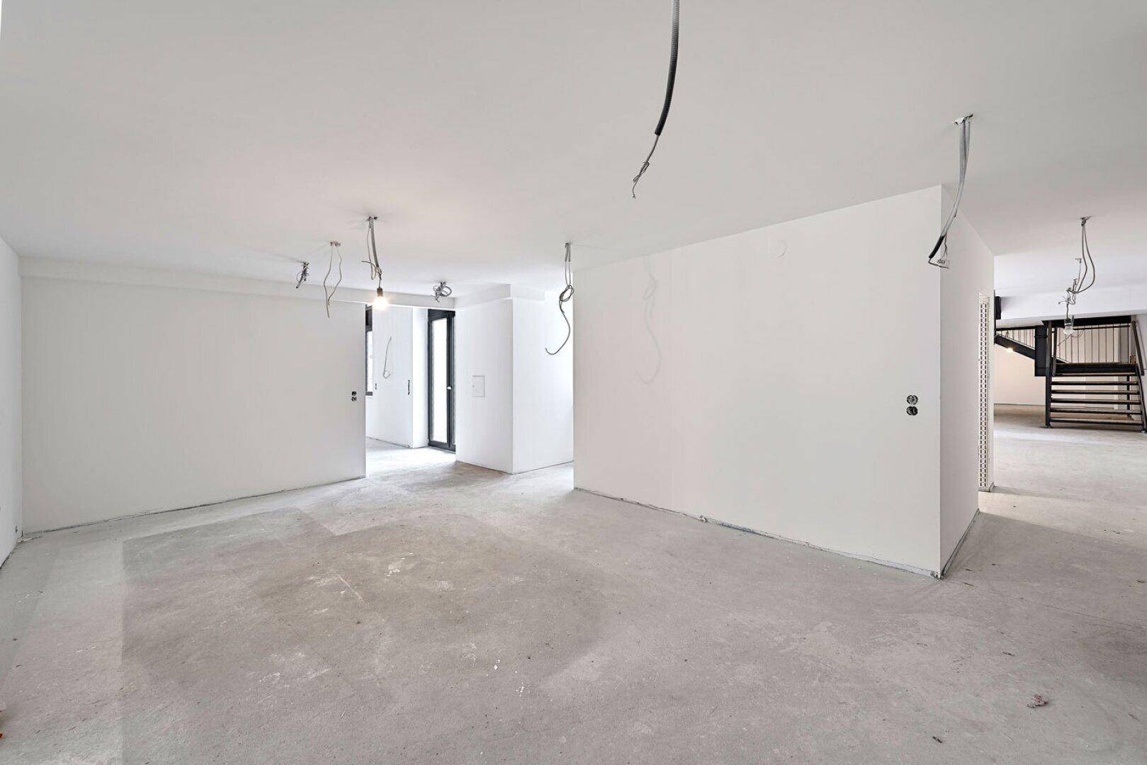 Leerer Büroraum im Rohbau mit weißen Wänden, Betonboden und sichtbaren Kabeln, bereit für den Ausbau.