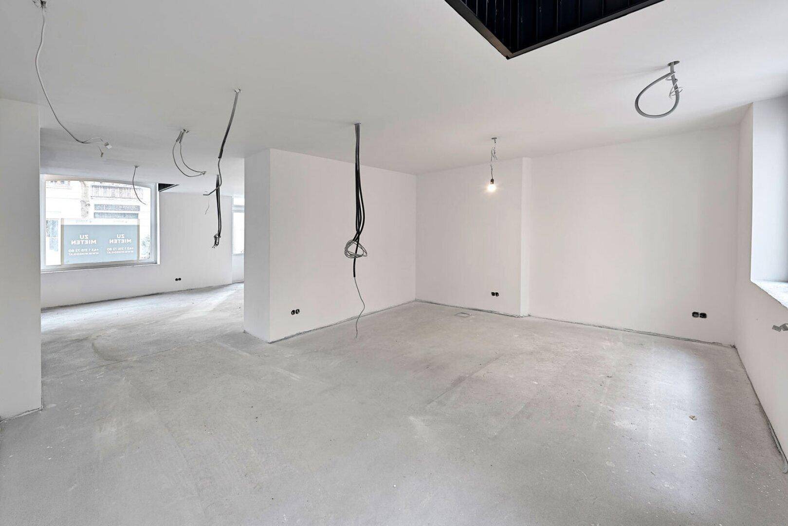 Großer, heller Büroraum im Rohbau mit Betonboden und freiliegenden Kabeln, Blick auf die Straße.