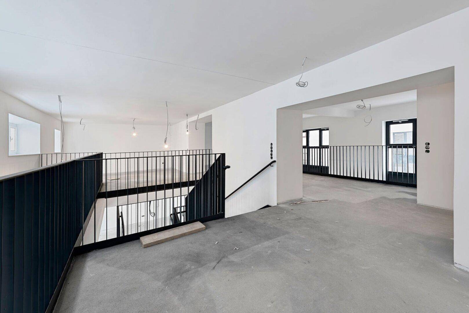 Offene Galerie in einem Büroraum im Rohbau mit schwarzem Geländer und Betonboden.