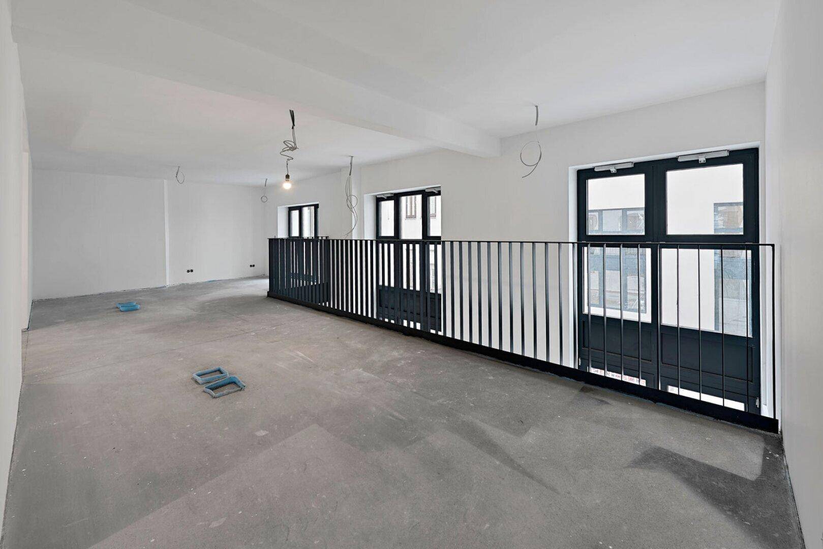 Langer Bürobereich im Rohbau mit schwarzem Geländer, Betonboden und großen Fenstern.
