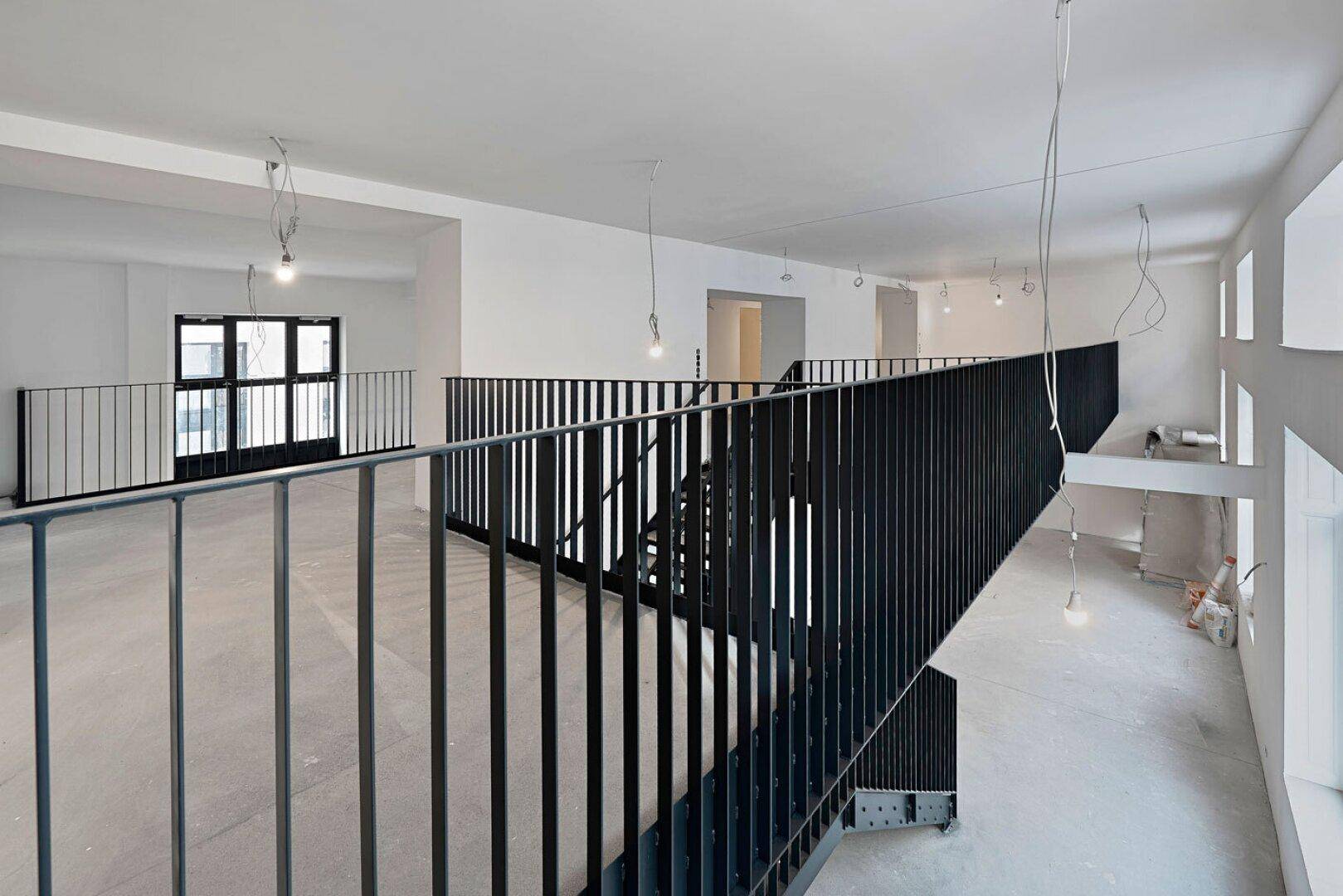 Blick von der Galerie in einen Büroraum im Rohbau mit Betonboden und freiliegenden Kabeln.