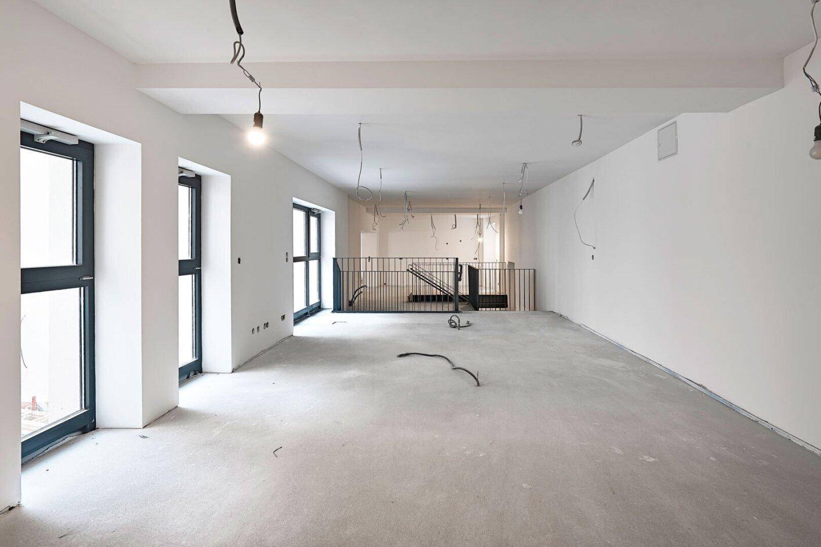 Großer, leerer Büroraum im Rohbau mit Betonboden, großen Fenstern und freiliegenden Kabeln.