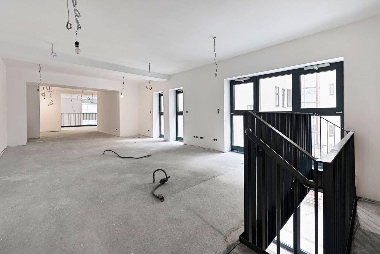 Leerer Büroraum im Rohbau mit Betonboden, großen Fenstern und einer schwarzen Metalltreppe.