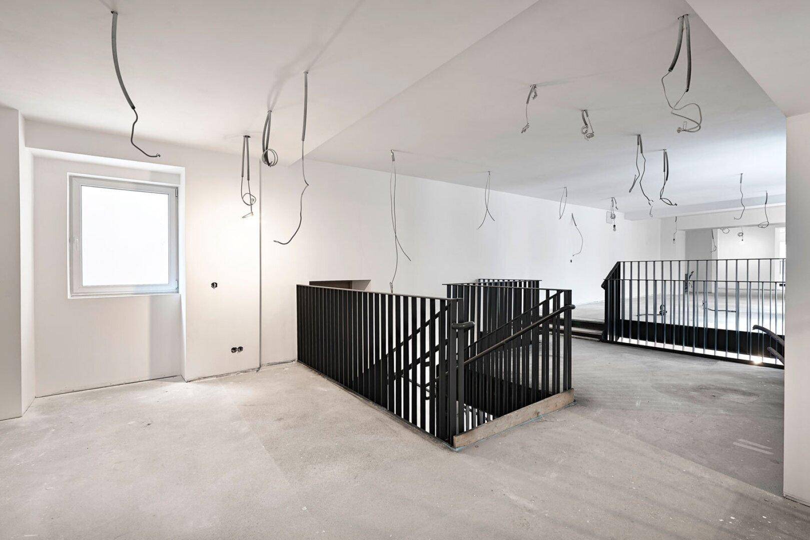 Bürobereich im Rohbau mit schwarzer Metalltreppe, Betonboden und freiliegenden Kabeln.