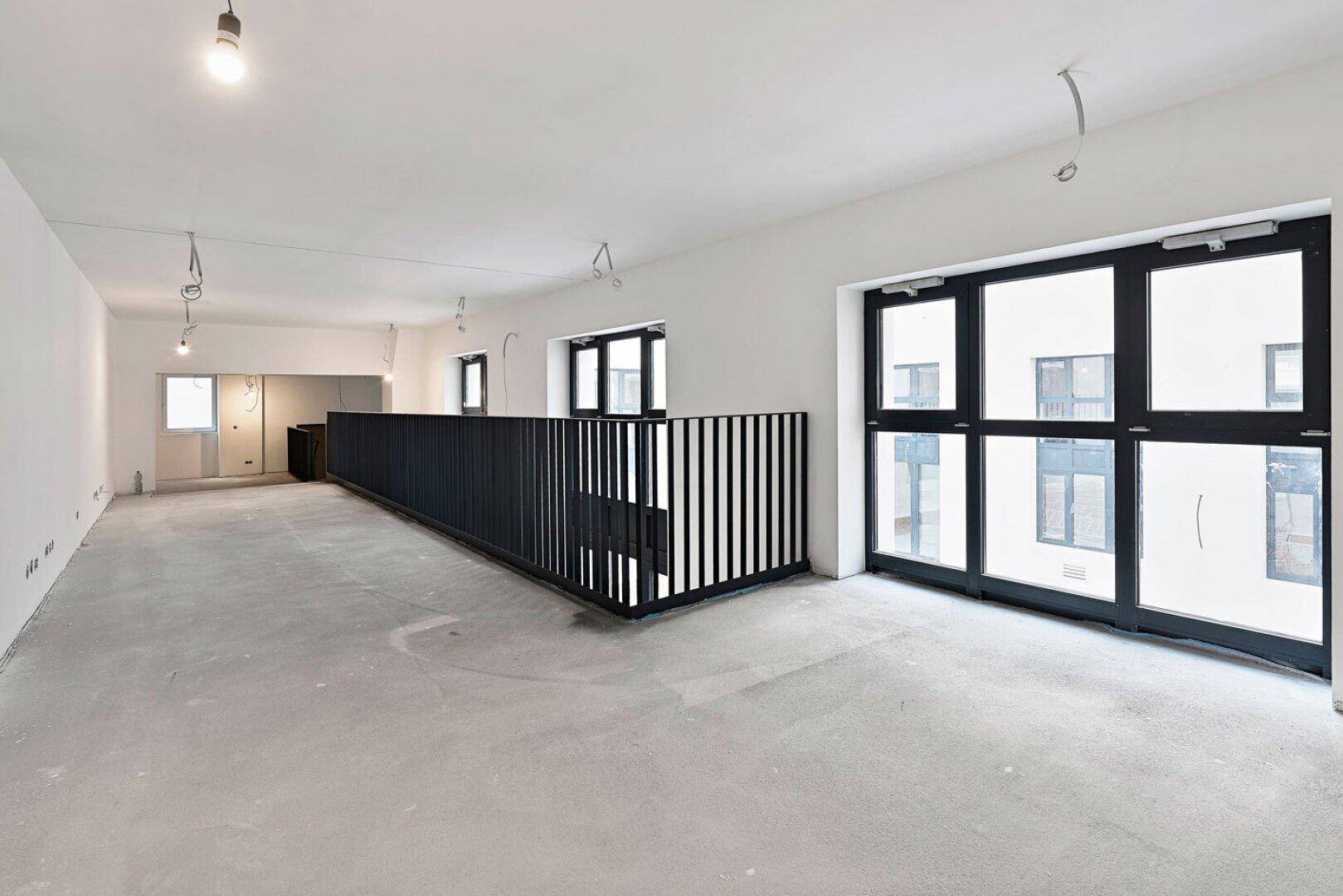 Langer Bürobereich im Rohbau mit schwarzem Geländer, Betonboden und großen Fenstern.