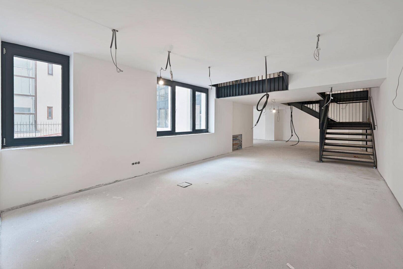 Großer, leerer Büroraum im Rohbau mit Betonboden, schwarzen Fenstern und freiliegenden Kabeln.