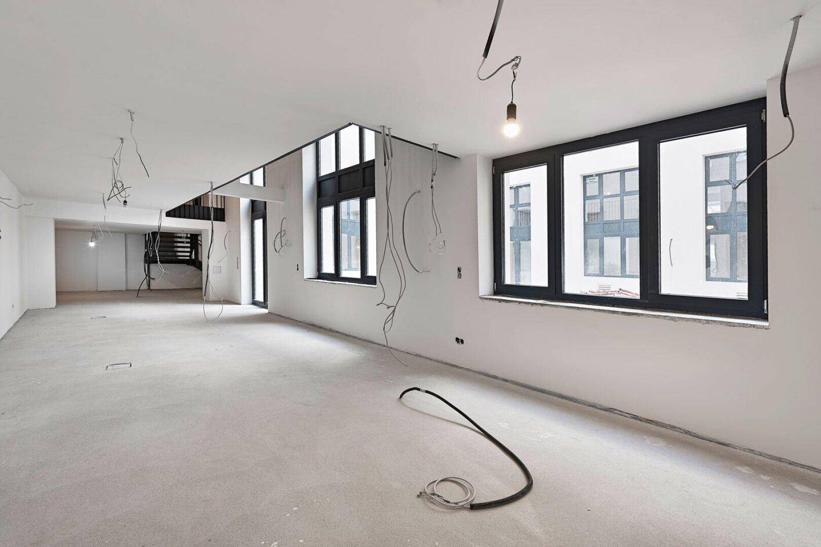 Weitläufiger Bürobereich im Rohbau mit Betonboden, schwarzen Fenstern und freiliegenden Kabeln.