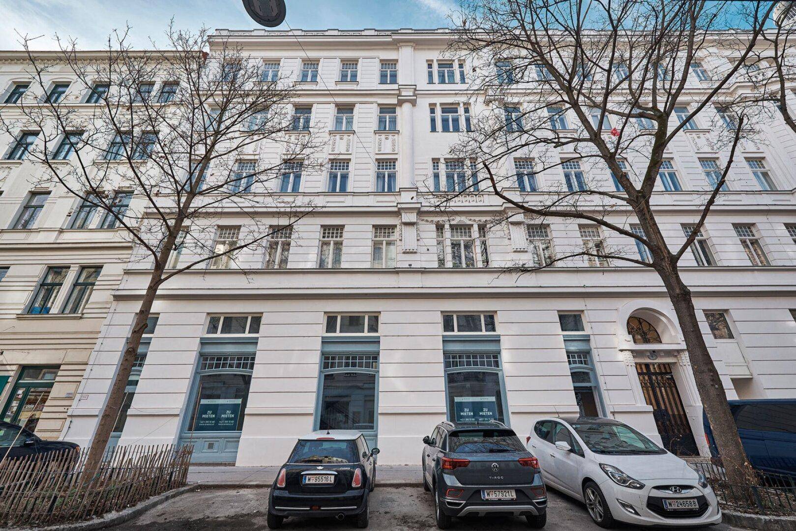 Historische Fassade eines Gebäudes mit vielen Fenstern und parkenden Autos davor.