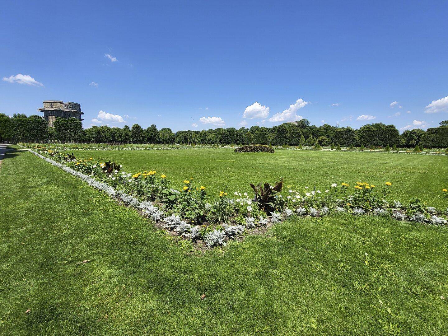 Weitläufige Grünfläche mit Blumenbeeten und einem Flakturm im Hintergrund unter blauem Himmel.