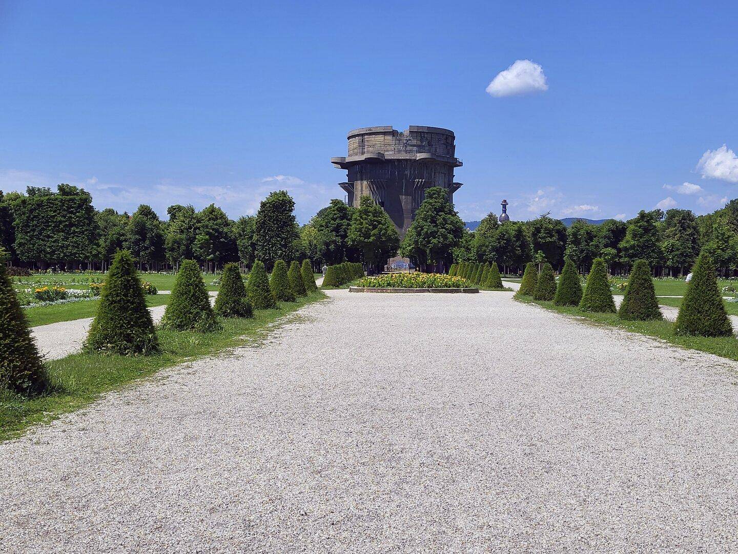 Kiesweg führt durch eine gepflegte Parkanlage mit Bäumen und einem Flakturm im Hintergrund.
