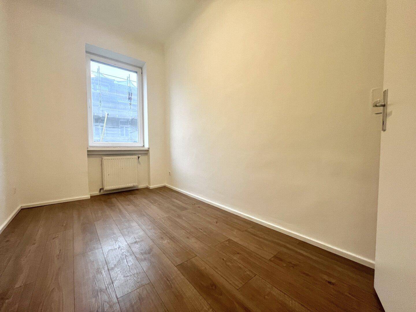 Kleines, helles Zimmer mit Holzboden und einem Fenster, das Tageslicht hereinlässt.