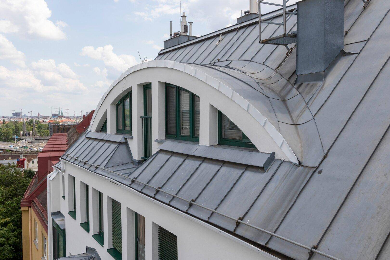 Detailansicht eines markanten Daches mit geschwungener Gaube und Metallverkleidung, die dem Gebäude Charakter verleiht.