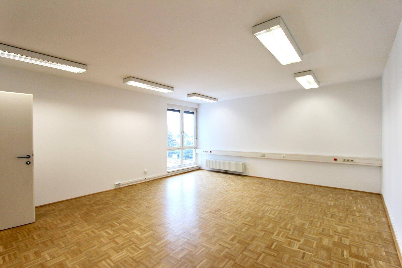Helles, leeres Büro mit Parkettboden, großen Fenstern und Deckenleuchten.